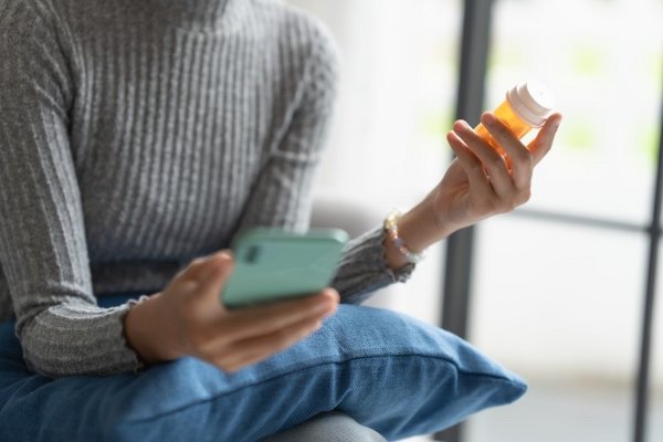 Eine Frau hält in der einen Hand ein Smartphone, in der anderen eine Dose mit Medikamenten.